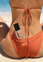 ZAYNA Fresh Blossom Face Mist tucked into the waistband of orange bikini bottoms on a sandy beach